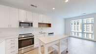 Located at 1200 N Henry St in Alexandria, VA, this well-appointed one-bedroom, one-bathroom unit offers a thoughtfully designed layout with modern finishes throughout. The open-concept kitchen and living area is anchored by a sleek, contemporary kitchen featuring white shaker cabinetry, light quartz countertops, a white subway tile backsplash with a warm wood accent niche, and a full suite of stainless steel appliances including a range, microwave, dishwasher, and refrigerator. Light wood-tone flooring runs throughout the main living space, creating a bright and cohesive feel, while recessed lighting ensures the area is well-illuminated. Large windows allow natural light to flow in, offering views of the surrounding Alexandria cityscape. The bedroom is carpeted for comfort and features a generously sized window with blinds that frame urban views, complemented by ample closet space — with four closets total throughout the unit. The bathroom is finished with large-format neutral tile in the tub/shower surround, paired with clean white walls and wood-tone flooring for a calm, spa-like aesthetic. The unit's clean, minimalist design and practical layout make it an efficient and stylish home in the heart of Alexandria.