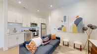 This thoughtfully designed single-floor unit offers a clean, contemporary aesthetic throughout, highlighted by bright white walls, recessed lighting, and light-toned hardwood flooring that unifies the open living spaces. The kitchen is outfitted with white shaker-style cabinetry featuring gold hardware, a white quartz countertop, a subway tile backsplash, and a full suite of stainless steel appliances including a range, microwave, dishwasher, and refrigerator. The kitchen flows seamlessly into the living area, creating an open-concept layout that maximizes the sense of space. The living area benefits from natural light streaming through large windows near the entry, contributing to an airy and welcoming atmosphere, complemented by colorful modern artwork on the walls. The bedroom is a comfortable retreat featuring plush carpeting, dual windows with roller shades that offer outdoor views, and access to a closet. The bathroom continues the unit's refined design language with a white vanity, quartz countertop, gold hardware and towel ring, an undermount sink, and a large wall mirror that enhances the sense of space. A hallway with a louvered closet door connects the living areas to the private spaces, maintaining an efficient and well-organized floor plan throughout.
