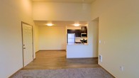 This single-floor space offers a thoughtfully designed open-concept layout that maximizes functionality and flow. The kitchen is a standout feature, boasting dark espresso cabinetry that provides a striking contrast against the warm yellow walls. It is fully equipped with stainless steel appliances, including a refrigerator, microwave, and electric range, complemented by a clean white countertop that wraps around a peninsula — ideal for casual dining or meal prep. The kitchen opens directly to the main living area, creating a seamless and connected atmosphere throughout the space. The living area features warm hardwood-style flooring that transitions to plush carpeting, adding comfort underfoot. Recessed flush-mount lighting illuminates the interior evenly, enhancing the welcoming ambiance. The walls are finished in a soft, warm yellow tone that lends a cozy and inviting feel to the entire space. A hallway near the entry extends the layout further, offering additional functional square footage. Overall, the space presents a clean, modern aesthetic with practical design choices that make it both livable and visually appealing.