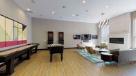 This thoughtfully designed single-floor amenity space offers a dynamic blend of recreation, socializing, and culinary functionality. The open-concept layout flows seamlessly between distinct zones, each serving a unique purpose while maintaining a cohesive contemporary aesthetic throughout.

The game room area is a standout feature, equipped with a foosball table, dual electronic dartboard machines, and a shuffleboard table, all set atop warm hardwood-style flooring. A wall-mounted flat-screen TV with a soundbar anchors the entertainment side of the room, complemented by stylish multi-armed pendant lighting overhead and a sleek linear fireplace set against a tiled accent wall.

Adjacent to the game room, a comfortable lounge area features patterned accent chairs arranged around low-profile tables atop a vibrant teal area rug, creating an inviting space to relax or socialize. A separate intimate sitting nook includes grey sofas, a bold yellow accent chair, and a large colorful abstract artwork, illuminated by a circular modern light fixture.

The fully equipped kitchen is bright and functional, showcasing dark cabinetry contrasted by white upper cabinets and quartz countertops, a subway tile backsplash, stainless steel appliances, and a large island with bar seating. A expansive window above the counter floods the kitchen with natural light and offers views of an outdoor courtyard.