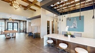 This thoughtfully designed community clubhouse spans a generous single floor, offering a seamless blend of modern elegance and inviting warmth throughout its open-concept layout. The entry reception area makes an immediate impression with dramatic exposed wood beam ceiling details, large-format tile flooring, sleek pendant lighting, and a white quartz-topped counter with stylish bar stools — setting a sophisticated tone from the moment guests arrive.

The space flows naturally into multiple distinct lounge areas, each curated with purposeful furnishings. One seating nook features light wood wall paneling, a living moss wall accent, and a mounted flat-screen TV, creating a cozy media retreat. A grand gathering room anchors the interior with soaring ceilings, a floor-to-ceiling stone fireplace surround, a linear electric firebox, exposed wood ceiling beams, an elegant chandelier, and an abundance of natural light pouring in through clerestory windows — all complemented by rich hardwood flooring and bold accent furnishings in warm orange and neutral tones.

A dedicated games area showcases a full-size billiards table beneath a statement lantern pendant, with access to a coffee and refreshment station nearby. Additional seating nooks with floor-to-ceiling patterned drapes and views of the surrounding outdoor pool area round out this versatile, amenity-rich communal space designed for both relaxation and social engagement.