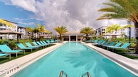 This outdoor amenity area centers around a generously sized rectangular pool with crystal-clear turquoise water, designed to evoke a resort-like atmosphere. The pool deck is finished in clean, light-toned concrete and lined symmetrically on both sides with rows of teal-cushioned chaise lounges and sleek metal-framed sunbeds, offering ample space for relaxation and sunbathing. Tall, mature palm trees are strategically positioned throughout the deck, providing natural shade and reinforcing the tropical aesthetic. At the far end of the pool, a structured cabana pavilion with a flat roof serves as a shaded gathering and lounge area, flanked by additional seating and umbrella-covered tables. The surrounding landscaping includes lush green turf accents that soften the hardscape and add visual warmth. The backdrop reveals a modern multi-story residential building with a contemporary facade, situating this amenity within a well-appointed community. The open sky above allows for abundant natural light throughout the day, enhancing the bright, airy ambiance. Overall, the space is thoughtfully laid out to balance functionality with an inviting, upscale outdoor living experience.