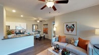 This single-story residence showcases an open-concept layout that effortlessly connects the living, dining, and kitchen areas into one cohesive living space. The living area is anchored by a comfortable neutral-toned sofa accented with warm coral and patterned throw pillows, complemented by a gold-framed coffee table and decorative wall art featuring a large butterfly motif. Ceiling fans are installed throughout, providing both functional airflow and a design element that enhances the airy feel of the space.

The kitchen features white cabinetry and a dark countertop peninsula that doubles as a breakfast bar, offering a natural visual boundary between the cooking and living zones without sacrificing the open feel. Adjacent to the kitchen, the dining area accommodates a round table with seating for multiple guests, positioned beneath a second ceiling fan. Dark hardwood-style flooring runs throughout, providing a rich contrast to the light-colored walls and white trim. Crown molding adds a refined architectural detail along the ceiling line. The overall ambiance is modern, welcoming, and well-appointed, with warm lighting enhancing the neutral color palette.