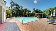 This outdoor pool area offers a well-designed recreational retreat set within a richly landscaped environment. The centerpiece is a generously sized rectangular swimming pool with clear turquoise water, framed by a classic brick-red paver deck that provides ample space for lounging and movement around the pool's perimeter. Stainless steel entry rails offer safe and easy access into the water.

A wooden pergola positioned along one side of the deck serves as a shaded gathering spot, adding architectural character to the outdoor setting. Lounge chairs and patio umbrellas are arranged around the pool, providing comfortable options for sun and shade. A compact utility shed is situated at one end of the pool area, housing what appears to be pool equipment and storage.

The entire space is enclosed by a black metal security fence with a gated entry, which features posted pool rules and safety signage. Blooming crepe myrtles and manicured shrubs add seasonal color, while tall, mature pine and deciduous trees form a dense natural canopy that borders the perimeter, offering privacy and a serene, park-like ambiance. The combination of structured hardscaping and abundant natural greenery creates an inviting and functional outdoor aquatic facility.