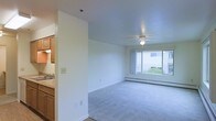This single-floor space is designed with a straightforward, functional layout that maximizes natural light and comfort. The main living area is carpeted and generously sized, featuring a ceiling fan with a light fixture overhead and a baseboard heating system running along the walls — practical elements that support year-round comfort.

Large windows are a defining feature of the space, with multiple panes positioned to capture outdoor views of mature trees and the surrounding neighborhood. The abundance of glazing ensures the interior remains bright throughout the day, creating a welcoming and open atmosphere.

The living area transitions seamlessly toward a kitchen space, partially visible through a dividing wall. The kitchen features warm wood-toned cabinetry and light countertops, and is finished with vinyl flooring that contrasts with the carpeted living area. A window in this area continues the theme of natural light throughout.

The neutral color palette — white and cream walls paired with light grey carpet — gives the space a clean, understated aesthetic that is easy to personalize. Overall, the space presents a functional and light-filled environment suited for everyday living.