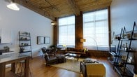 This industrial-style loft seamlessly blends raw architectural character with modern comfort across a single, expansive floor. The living area commands immediate attention with its dramatically high ceilings, where original exposed timber beams and ceiling joists run the length of the space, lending a warm, warehouse aesthetic. Original brick walls frame the perimeter, adding texture and historic charm to the interior. Oversized windows stretch nearly floor to ceiling, allowing abundant natural light to pour in and illuminate the rich hardwood floors below. The open-plan living area is thoughtfully arranged with mid-century modern furnishings, including a tufted sofa, lounge chairs, and a central coffee table set atop a natural fiber rug, creating an inviting and cohesive gathering space. A separate seating nook near the windows provides an intimate corner for conversation or quiet reading. Industrial pendant lighting and arc floor lamps complement the space's character while providing warm, layered illumination throughout. The interplay of reclaimed wood, exposed brick, gleaming hardwood floors, and expansive glazing creates a dynamic ambiance that is both urban and deeply welcoming — a refined take on classic loft living.