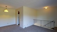 This single-floor space presents a clean, straightforward layout with neutral cream-colored walls and wall-to-wall grey carpeting that contributes to a simple, understated aesthetic. A textured ceiling adds subtle visual interest overhead. One of the more distinctive architectural features is the wrought iron stair railing along one edge of the space, suggesting an open upper-level landing that overlooks a lower level, providing a sense of vertical openness despite the single-floor designation. A partial wall divides the main open area from an adjacent kitchen, which is visible through a doorway and includes a white refrigerator. The transition between spaces is defined by clean white trim and baseboards that frame the walls consistently throughout. The overall ambiance is neutral and functional, with ample wall space and an open feel that lends itself well to flexible furniture arrangements. The space is well-suited for those seeking a straightforward, low-maintenance environment with a practical and understated design.