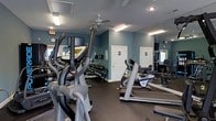 Located at 2890 Woodland Dr in New Bern, North Carolina, this fitness center offers a comprehensive workout facility designed to support a wide range of exercise routines. The space is laid out on a single floor and features a dark rubber flooring surface ideal for high-traffic gym use. The interior is painted in a calming blue-gray tone, contributing to a focused and energizing atmosphere.

The facility is well-equipped with a variety of cardio machines, including treadmills, elliptical trainers, and a stair climber, providing ample options for aerobic exercise. A multi-station cable and weight machine serves as the centerpiece for strength training, complemented by a free-weight dumbbell rack along the wall. Large mirrors line the walls, visually expanding the space and allowing users to monitor their form during workouts.

Overhead ceiling fans and fluorescent lighting ensure the space remains well-ventilated and brightly lit. Restroom facilities are conveniently accessible directly from the gym floor, and a vending machine is available near the entrance. A wall clock keeps members on schedule throughout their sessions. The overall layout is open and functional, making efficient use of the available floor space to accommodate multiple users simultaneously.