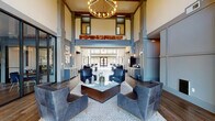 Located at 950 Riverside Dr in Chattanooga, Tennessee, this sophisticated clubhouse serves as a premier amenity space defined by its striking architectural design and refined interior aesthetic. The grand entrance lobby immediately commands attention with its dramatic double-height ceilings, structural columns clad in slate-blue paneling, and a statement globe chandelier that anchors the space. A large arched window at the rear floods the room with natural light and frames outdoor views, while rich hardwood flooring in a herringbone-laid pattern extends throughout. The main lounge area is appointed with plush navy and charcoal leather seating arranged around a glass-topped coffee table, creating an inviting setting for gathering or relaxing. Adjacent to the lobby, a dedicated coffee bar station offers a convenient self-serve beverage area with custom cabinetry finished in deep gray with gold hardware. Further into the space, a secondary lounge and dining area features velvet blue chairs with brass-finished legs, warm wood tables, and curated contemporary artwork adorning the walls. Wall sconces provide warm ambient lighting throughout. A separate media lounge room, visible through a wide doorway, offers a more intimate retreat with dark-toned walls, built-in shelving, a large television, and eclectic seating. The overall ambiance blends modern sophistication with comfortable, community-focused design.