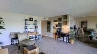 This single-floor space showcases an open-concept layout that seamlessly connects the living area to the kitchen. The living area is finished with wall-to-wall carpet, lending a warm and comfortable feel underfoot. A metal-framed shelving unit lines one wall, offering display space for decorative items and personal belongings, while a seating arrangement with an ottoman adds to the room's practical comfort.

A dedicated workspace is positioned toward the center of the room, featuring a dark wood desk and chair, making the space well-suited for both relaxation and productivity. Decorative wall art panels are arranged above and around the pass-through area, adding visual interest and a curated aesthetic to the neutral-toned walls.

The kitchen, visible through an open passageway, features light wood cabinetry and appears bright and functional. A tall mirror near the kitchen entry enhances the sense of depth in the space. Potted plants introduce a touch of greenery, complementing the warm, neutral palette. The textured ceiling and consistent use of soft, natural tones contribute to an overall calm and cohesive ambiance throughout.