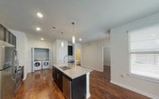 This single-story residence at 401 S Coit in McKinney, Texas offers a well-designed, open-concept layout with rich, dark hardwood flooring throughout. The kitchen serves as the centerpiece of the home, boasting a large granite-topped island with a stainless steel undermount sink and pendant lighting above. Dark espresso cabinetry lines the walls and pairs elegantly with stainless steel appliances, including a refrigerator, range, and microwave. A tile backsplash adds a clean, polished finish to the cooking area. The open layout flows seamlessly into the living area, which is bright and airy, illuminated by recessed lighting and a ceiling fan. The bedrooms continue the warm hardwood flooring and feature ceiling fans, ample natural light through blinds-covered windows, and generous closet space. The bathroom is well-appointed with large-format white tile surrounding a soaking tub and shower combination, dark vanity cabinetry, granite countertops, and a framed mirror. A dedicated laundry area accommodates a full-size side-by-side washer and dryer. The home's neutral palette and modern finishes create a clean, contemporary ambiance throughout.