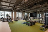 This fitness center combines industrial-modern design with a comprehensive range of workout amenities across a generously sized, single-level space. The exposed ceiling showcases visible ductwork, piping, and pendant lighting fixtures, lending the facility an urban, loft-like aesthetic. Large windows line multiple walls, flooding the interior with natural light while offering views of the surrounding cityscape.

The flooring is divided into two functional zones: a green rubberized turf area designated for functional training and a smooth neutral-toned surface accommodating cardio and strength equipment. The cardio section features a row of stationary cycling bikes and treadmills positioned near the windows. The strength and functional training area is equipped with dumbbell racks, medicine ball storage, TRX suspension trainers, plyo boxes, balance domes, a freestanding punching bag, and ab benches.

A large flat-screen display is mounted on one wall, suitable for guided workout sessions. Built-in storage cubbies and bench seating line the perimeter near the entrance corridor, which leads to additional rooms, likely changing or locker facilities. A glass door provides access to an outdoor area. The overall ambiance is energetic, modern, and well-organized for diverse training needs.