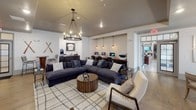 This thoughtfully designed single-floor amenity space blends comfort, functionality, and entertainment into a cohesive, modern farmhouse aesthetic. The open-concept lounge serves as the heart of the space, anchored by a generous sectional sofa, a rustic drum coffee table, and a large wall-mounted flat-screen TV. A hoop-style chandelier overhead adds an industrial warmth, while framed sports jerseys and memorabilia lend the space a spirited, personalized character. Warm hardwood-style flooring flows throughout, tying the zones together seamlessly.

A dedicated workstation nook lines one wall, featuring a built-in wooden desk with multiple computer stations illuminated by exposed Edison bulb pendant lights against a shiplap accent wall — offering a productive counterpoint to the leisure areas. A coffee and beverage station nearby adds everyday convenience.

Adjacent to the main lounge, a game room hosts a round poker table surrounded by upholstered chairs and bathed in natural light from large windows with elegant draping. Framed artwork decorates the walls, adding a polished finish. A full-length shuffleboard table occupies another zone, providing additional recreational options.

The overall ambiance is inviting and well-appointed, striking a balance between a relaxed gathering space and a fully functional community hub.