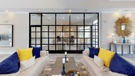 This thoughtfully designed single-floor amenity space combines modern aesthetics with functional comfort throughout its open, airy layout. The centerpiece of the main lounge is a soaring vaulted ceiling with a contemporary chandelier and a dramatic floor-to-ceiling black-framed glass partition wall, which visually connects the lounge to a private office suite while maintaining a sense of openness and transparency. Light hardwood flooring flows throughout, complementing the crisp white walls and neutral furnishings accented with bold navy and yellow throw pillows. Large black-framed glass entry doors and generous windows flood the interior with natural light, offering views of the surrounding greenery. A seating area near the entrance features plush armchairs arranged around a gold accent table, creating an inviting gathering spot. The kitchen is well-appointed with white shaker cabinetry, open floating shelves, white subway tile backsplash, stainless steel appliances including a full-size refrigerator and range hood, and a spacious marble-top island with bar seating. Adjacent to the main lounge, a cozy dining nook provides a round wooden table with upholstered chairs, ideal for casual meetings or dining. The overall ambiance is bright, contemporary, and welcoming.