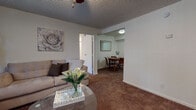 This single-floor residence offers a thoughtfully organized layout designed for comfortable everyday living. The living room serves as the heart of the home, featuring warm carpeted flooring, a ceiling fan, a textured ceiling, and a large window fitted with vertical blinds that allows natural light to filter in while providing a view of surrounding greenery. The space flows seamlessly into a defined dining area, which accommodates a round wooden table and chairs and is positioned adjacent to the kitchen.

The galley-style kitchen is efficiently designed with dark cabinetry, light granite-style countertops, a double-basin sink, and white appliances including a range and refrigerator. Wood-look flooring adds a clean, updated feel to this functional workspace.

Two bedrooms provide restful retreats, each with plush carpeting, ceiling fans, and windows with vertical blinds that bring in soft natural light. One bedroom features a dark-toned upholstered headboard with matching nightstands, while the second offers a warm wood bedroom set with a mirrored dresser.

The bathrooms are well-appointed, featuring dark wood vanities with granite-style countertops, wood-look tile flooring, and clean, bright fixtures. A carpeted hallway lined with closet doors connects the private areas of the home, adding ample storage throughout.