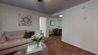 This single-floor residence offers a thoughtfully organized layout designed for comfortable everyday living. The living room serves as the heart of the home, featuring warm carpeted flooring, a ceiling fan, a textured ceiling, and a large window fitted with vertical blinds that allows natural light to filter in while providing a view of surrounding greenery. The space flows seamlessly into a defined dining area, which accommodates a round wooden table and chairs and is positioned adjacent to the kitchen.

The galley-style kitchen is efficiently designed with dark cabinetry, light granite-style countertops, a double-basin sink, and white appliances including a range and refrigerator. Wood-look flooring adds a clean, updated feel to this functional workspace.

Two bedrooms provide restful retreats, each with plush carpeting, ceiling fans, and windows with vertical blinds that bring in soft natural light. One bedroom features a dark-toned upholstered headboard with matching nightstands, while the second offers a warm wood bedroom set with a mirrored dresser.

The bathrooms are well-appointed, featuring dark wood vanities with granite-style countertops, wood-look tile flooring, and clean, bright fixtures. A carpeted hallway lined with closet doors connects the private areas of the home, adding ample storage throughout.