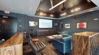 Located at 55 E 9th Ave in Columbus, Ohio, this single-floor space presents a thoughtfully designed interior with a distinctly modern and upscale aesthetic. The centerpiece of the space is a dedicated screening or presentation lounge, anchored by a large projection screen mounted along one wall, complemented by an additional flat-panel display nearby. Dark, moody tones define the room, with deep charcoal-gray walls and a dark ceiling panel that houses a grid of directional track lighting and a mounted projector, creating a cinematic ambiance. The seating arrangement is curated and stylish, featuring tufted black leather lounge chairs paired with a round wooden side table, alongside plush teal velvet sofas arranged around a sleek glass-and-metal coffee table. A low media credenza runs beneath the screens, providing both functional storage and a clean visual base. Framed artwork on the walls adds personality and a gallery-like quality to the space. The overall atmosphere is polished and intimate, blending the functionality of a professional presentation environment with the comfort and design sensibility of a high-end lounge.