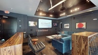 Located at 55 E 9th Ave in Columbus, Ohio, this single-floor space presents a thoughtfully designed interior with a distinctly modern and upscale aesthetic. The centerpiece of the space is a dedicated screening or presentation lounge, anchored by a large projection screen mounted along one wall, complemented by an additional flat-panel display nearby. Dark, moody tones define the room, with deep charcoal-gray walls and a dark ceiling panel that houses a grid of directional track lighting and a mounted projector, creating a cinematic ambiance. The seating arrangement is curated and stylish, featuring tufted black leather lounge chairs paired with a round wooden side table, alongside plush teal velvet sofas arranged around a sleek glass-and-metal coffee table. A low media credenza runs beneath the screens, providing both functional storage and a clean visual base. Framed artwork on the walls adds personality and a gallery-like quality to the space. The overall atmosphere is polished and intimate, blending the functionality of a professional presentation environment with the comfort and design sensibility of a high-end lounge.