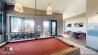 Located in the heart of downtown Cary, NC on E Chatham Street, this contemporary amenity lounge offers a well-appointed, single-floor space thoughtfully divided into distinct functional areas. The interior features soaring ceilings and an open layout that fosters a welcoming social atmosphere. A standout billiards room anchors one side of the space, complete with a red felt pool table, wall-mounted smart TV, built-in wet bar, pendant lighting, and large floor-to-ceiling windows that flood the room with natural light and frame treetop views of the surrounding area. Adjacent to this is a comfortable lounge area furnished with a curved sectional sofa, accent chairs, and a distinctive live-edge wood coffee table, complemented by built-in shelving and an additional wall-mounted display. A dining nook with a round table and upholstered chairs provides a more intimate gathering spot. Large glass sliding doors seamlessly connect the interior to a covered outdoor terrace, which features a warm wood-paneled ceiling, ceiling fans, and open-air seating with woven accent chairs and café-style tables. The terrace extends to an open balcony with mesh railings, offering sweeping views of the Cary skyline and surrounding greenery—an ideal setting for relaxation or social gatherings in any season.