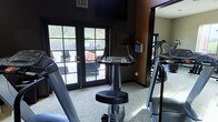 This single-level fitness center offers a focused workout environment equipped with essential cardio machines, including a stationary bike, treadmill, and elliptical trainer. The room features dark-painted walls that create a sleek, modern atmosphere, complemented by neutral carpeted flooring. A large mirror along one wall visually expands the space and allows users to monitor their form during exercise. Natural light filters in through a set of classic French doors, which open directly to an outdoor patio or pool area, providing pleasant views of surrounding greenery and hills. A wall-mounted flat-screen television is positioned for viewing during workouts, adding an entertainment element to the space. Posted fitness center guidelines are displayed on the wall, keeping users informed of facility rules. The combination of natural light from the glass-paned doors and the dark interior palette gives the room a balanced, energizing ambiance. The layout is efficiently organized to maximize usable floor space while keeping equipment accessible and the overall environment comfortable for daily exercise routines.