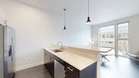 This efficiently designed one-bedroom, one-bathroom unit offers a contemporary urban living experience spread across a single floor. The open-concept kitchen and living area serves as the heart of the space, featuring dark hardwood flooring, crisp white walls, and large windows that invite natural light and provide views of the surrounding neighborhood. The kitchen is outfitted with stainless steel appliances, including a range, over-the-range microwave, and refrigerator, complemented by dark cabinetry, white quartz countertops, and a peninsula with pendant lighting overhead — ideal for casual dining or meal prep. The living area offers a comfortable, minimalist layout with exposed metallic ductwork running along the ceiling, lending an industrial edge to the otherwise clean aesthetic. A long, well-lit hallway with a sliding barn-style door connects the main living area to the private bedroom and bathroom. The bedroom is a cozy, carpeted retreat featuring a sliding closet with built-in shelving and recessed lighting above. The bathroom maintains the unit's modern design language with a dark floating vanity, white quartz countertop, full-width mirror, light tile flooring, and chrome fixtures throughout.