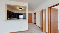 This single-floor space is thoughtfully laid out to maximize functionality and flow. The kitchen is framed by a built-in pass-through window that opens to the main living area, creating an open, connected feel. Dark cabinetry with a contrasting light countertop lines the kitchen walls, complemented by stainless steel appliances and recessed overhead lighting, giving the cooking area a clean, modern character.

The living area transitions seamlessly from the kitchen via a carpeted floor that extends throughout the main hallway and adjoining rooms, adding warmth underfoot. Neutral white walls and light-toned baseboards and door trim maintain a bright, airy atmosphere throughout.

Down the hallway, a utility area is visible, housing what appears to be a stacked washer and dryer unit — a practical in-unit feature. Adjacent to this, a sliding door reveals a spacious closet, offering generous storage. The front entry door, finished in warm wood tones, anchors the end of the hallway and provides a welcoming focal point. Recessed ceiling lighting runs through the corridor, ensuring the space remains well-lit throughout.