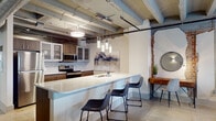 This single-floor urban loft masterfully balances industrial heritage with contemporary design. Throughout the space, polished terrazzo-style concrete floors, exposed structural beams, and open ductwork celebrate the building's raw architectural character, while modern finishes bring warmth and refinement.

The open-concept kitchen is a standout feature, equipped with stainless steel appliances, wood-toned cabinetry with glass-front upper panels, a mosaic tile backsplash, and a generous white countertop island with bar seating and pendant lighting overhead. A dining area anchored by a round wood table sits alongside an original exposed brick wall, adding texture and historic charm.

The living area offers a comfortable, well-lit retreat with a large window framing urban views and exposed ductwork above that reinforces the loft's industrial aesthetic. The bedroom continues the cohesive design with polished floors, exposed brick accents, large windows with roller shades, and direct access to a private en-suite bathroom featuring a backlit mirror, wood vanity, subway tile walls, and a shower-tub combination. A second full bathroom mirrors this clean, modern styling. Together, the spaces create a cohesive, urban living environment rich in character and detail.