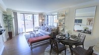 This single-floor residence presents an open-concept layout that effortlessly connects the living and dining areas, creating a cohesive and inviting atmosphere. The living area is anchored by a plush, light-toned sofa accented with patterned throw pillows, complemented by a sleek glass coffee table and a warm terracotta upholstered bench that adds a striking pop of color. A large sliding glass door floods the space with natural light and opens directly onto a private balcony, offering views of the surrounding neighborhood. Hardwood flooring runs throughout, lending warmth and continuity to the space. A stylish open metal shelving unit serves as a functional divider between the living and dining zones, displaying decorative bottles, glassware, and curated accessories. The dining area features a round dark-wood table paired with classic wooden chairs, set with elegant place settings and a fresh floral centerpiece. Wall-mounted sconces and coordinated floor lamps provide warm ambient lighting throughout. The overall ambiance is modern yet inviting, with a sophisticated blend of neutral tones, layered textures, and carefully chosen furnishings that create a comfortable and visually refined living environment.