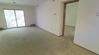 This single-floor space presents a straightforward and functional layout, characterized by open, generously sized rooms connected through a wide doorway. The interior is finished with wall-to-wall carpeting in a neutral beige-gray tone, providing a soft, uniform foundation throughout. The walls are painted in a clean, off-white palette, contributing to a bright and airy atmosphere.

Natural light enters through a window positioned along one wall, adding warmth to the open living area. A baseboard heating unit is visible along the perimeter, offering an efficient heating solution integrated neatly into the room's design. The trim and door frames are finished in warm wood tones, providing a subtle contrast against the lighter walls and flooring.

The adjoining room, visible through the wide doorway, mirrors the same carpeting and wall finish, suggesting a consistent design language carried throughout the space. Electrical outlets are distributed along the walls, and a wall-mounted light fixture is present overhead. The overall ambiance is clean, neutral, and move-in ready, offering a blank canvas suitable for a variety of uses and furnishing styles.