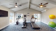This fitness center is housed within a single-floor space defined by striking vaulted ceilings with white-painted exposed beams and tongue-and-groove paneling, creating an airy, open atmosphere. Multiple ceiling fans are mounted along the roofline, ensuring consistent airflow throughout the workout area. Large sliding glass doors and windows line multiple walls, flooding the interior with natural light and providing views of the surrounding outdoor landscape.

The flooring throughout is a patterned, blue-toned carpet that adds both comfort and visual interest underfoot. The cardio zone is well-appointed with a treadmill, elliptical trainer, and a stationary spin bike, all positioned to take advantage of the natural light and outdoor views. A dedicated strength training area features a full cable and pulley multi-station machine, a weight bench, and a tiered dumbbell rack stocked with a full range of free weights.

Accent walls clad in vertical wood slat paneling add a warm, modern design element that contrasts elegantly with the white ceiling and neutral walls. A wall-mounted flat-screen television is positioned in the strength training zone for entertainment during workouts. The overall ambiance is bright, clean, and inviting, combining functional fitness amenities with thoughtful architectural detailing.