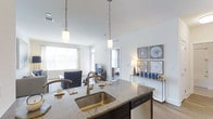 Located at Kensington Place, this single-story residence offers a thoughtfully designed open-concept layout that connects the kitchen and living areas in a bright, cohesive space. The kitchen serves as a central hub, featuring a large island with granite countertops, a deep stainless steel sink, and pendant lighting with warm brass accents that hang from soaring ceilings. A dishwasher is integrated beneath the island, adding functional convenience. The open flow leads directly into the living area, which is filled with abundant natural light streaming through large windows. Walls are finished in crisp white, enhancing the airy, clean ambiance throughout. Hardwood-style flooring runs continuously across the space, lending a polished, cohesive look. Decorative wall art adds a curated touch to the living area, while a glimpse into the adjacent bedroom reveals a similarly bright and well-appointed room. The high ceilings amplify the sense of openness, making the overall space feel expansive and welcoming. The layout and finishes combine to create a modern, comfortable living environment.