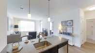 Located at Kensington Place, this single-story residence offers a thoughtfully designed open-concept layout that connects the kitchen and living areas in a bright, cohesive space. The kitchen serves as a central hub, featuring a large island with granite countertops, a deep stainless steel sink, and pendant lighting with warm brass accents that hang from soaring ceilings. A dishwasher is integrated beneath the island, adding functional convenience. The open flow leads directly into the living area, which is filled with abundant natural light streaming through large windows. Walls are finished in crisp white, enhancing the airy, clean ambiance throughout. Hardwood-style flooring runs continuously across the space, lending a polished, cohesive look. Decorative wall art adds a curated touch to the living area, while a glimpse into the adjacent bedroom reveals a similarly bright and well-appointed room. The high ceilings amplify the sense of openness, making the overall space feel expansive and welcoming. The layout and finishes combine to create a modern, comfortable living environment.