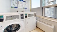 Located at 100 Roehampton Ave in Toronto's Midtown neighbourhood, this shared laundry facility offers a compact, functional space designed to serve building residents efficiently. The room is outfitted with commercial-grade Huebsch coin-operated washing machines and dryers, managed through the Coinamatic service network, ensuring reliable and accessible laundry services. A white utility sink is positioned near the window, providing a convenient handwashing or pre-treatment station. The light-coloured tile flooring throughout the space is easy to maintain and contributes to the room's clean, utilitarian aesthetic. Neutral grey walls keep the environment bright and orderly. A window allows natural light to fill the space and offers a view of the surrounding urban skyline, featuring the modern high-rise buildings characteristic of the Midtown Toronto area. Instructional signage for washer and dryer operation is clearly posted on the walls, ensuring straightforward use for all residents. The entrance features a self-closing door with a posted reminder to keep it shut, supporting building security and noise control. Overall, this laundry room provides a practical, well-equipped shared amenity in a convenient Midtown Toronto location.