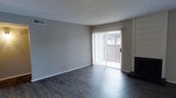 This single-story space features a clean, straightforward layout with practical finishes throughout. The main living area is anchored by dark hardwood-style flooring that contrasts with the neutral gray walls, creating a balanced, understated aesthetic. A textured popcorn ceiling adds a classic touch overhead, while white baseboards provide a crisp border along the floor.

Natural light enters generously through a large sliding glass door fitted with vertical blinds, which opens to an outdoor balcony enclosed by a wooden railing. This connection to the exterior brings a sense of openness to the interior and allows for easy indoor-outdoor flow.

A fireplace with a brick surround is positioned along one wall, serving as a functional focal point within the living area. The fireplace features a raised hearth and dark trim, adding a traditional element to the otherwise minimalist space.

The overall ambiance is calm and neutral, offering a blank-canvas quality that makes the space highly adaptable for various furnishing and décor styles. The combination of durable flooring, natural light, and the fireplace makes this a comfortable and functional living environment.