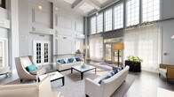 This sophisticated community space makes an immediate impression with its grand, rotunda-style foyer, where a sweeping double staircase with wrought-iron railings frames a striking circular dark wood table beneath an ornate chandelier. Stone tile flooring, marble-accented wall panels, and curated seating alcoves contribute to the refined, welcoming character of the entry. Towering ceilings with a dramatic arched transom window above the main entrance doors draw the eye upward and flood the space with natural light. Transitioning from the foyer, a spacious lounge area continues the sense of grandeur with soaring double-height ceilings adorned with coffered detailing and an expansive grid of oversized windows that bathe the interior in daylight and offer views of the surrounding greenery. White paneled walls, rich dark hardwood flooring, and a classic fireplace anchor the lounge, which is furnished with crisp white sofas and accent chairs accented by blue and teal throw pillows. French doors with glass panes connect the main lounge to additional rooms, creating an open and flowing layout. Throughout, the design palette of cool greys, soft blues, and warm neutrals maintains a cohesive, polished aesthetic that balances elegance with approachable comfort.