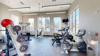 This fitness center offers a comprehensive workout environment designed to accommodate a wide range of exercise routines. The space is anchored by large, floor-to-ceiling windows along multiple walls, flooding the interior with abundant natural light and providing pleasant views of the surrounding trees and outdoor scenery. The durable rubber flooring throughout provides both safety and comfort underfoot.

The cardio zone is well-appointed with multiple treadmills, elliptical trainers, and a recumbent bike, all positioned to take advantage of the natural light and outdoor views. A wall-mounted television is conveniently located nearby for entertainment during workouts. The strength training area features a multi-station cable machine, a dedicated chest press machine, and an adjustable weight bench, catering to a variety of resistance training needs.

A dedicated free weights section includes a full rack of dumbbells, barbells with weight plates, and an extensive collection of kettlebells and medicine balls, supporting functional and dynamic training. Overhead fluorescent lighting supplements the natural light, ensuring the space remains bright throughout the day. The neutral warm-toned walls and open layout contribute to an inviting and energizing atmosphere, making this fitness center a practical and motivating facility for daily use.
