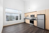 This well-designed single-floor residence offers a clean, contemporary aesthetic with thoughtful finishes throughout. The kitchen is a standout feature, equipped with light wood cabinetry extending to the ceiling, a full suite of stainless steel appliances including a refrigerator, electric range with overhead microwave, and dishwasher, all complemented by a gray countertop with an integrated sink. Durable hardwood-style flooring runs through the main living areas and kitchen, adding warmth and cohesion to the space. A bright hallway connects the rooms, featuring recessed lighting and a ceiling access panel, guiding residents toward the bedroom areas. One bedroom is finished with the same hardwood-style flooring and features tall windows that frame views of the surrounding residential neighborhood, allowing generous natural light to fill the room. A second bedroom offers plush carpeting underfoot and a sliding window with views of a covered outdoor area. The full bathroom is cleanly appointed with a white soaking tub and shower combo, chrome fixtures, and matching hardwood-style flooring. Neutral warm-toned walls throughout the space provide a versatile backdrop, while the overall layout offers a comfortable and practical living environment.