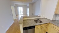 This single-floor residence offers an open-concept layout that seamlessly connects the kitchen and living areas, creating a welcoming and functional living environment. The kitchen is appointed with speckled granite countertops that extend across a peninsula, providing ample prep space and a casual dining or workspace area. A stainless steel sink and dishwasher are integrated into the peninsula, complementing the light-toned cabinetry beneath. The open layout allows for a clear sightline from the kitchen into the expansive living area, which features warm hardwood-style flooring that runs throughout. Large sliding glass doors at the far end of the living space flood the interior with natural light and provide direct access to a private balcony, offering an extension of the living space outdoors. The high ceilings contribute to an airy, spacious feel throughout the unit. Neutral wall tones provide a clean, versatile backdrop suitable for a variety of interior styles. Additional features include visible HVAC venting and ample electrical access points positioned conveniently throughout. The overall ambiance is bright, modern, and move-in ready, making it a practical and comfortable living space.