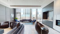 Located on N State St in Chicago's vibrant Gold Coast neighborhood, this elegantly appointed amenity floor delivers a refined blend of modern design and thoughtful functionality. The main lounge area is anchored by floor-to-ceiling windows that flood the space with natural light and frame striking views of the Chicago skyline. Curved brown leather chairs, a sleek gray sofa, and blue accent seating are arranged around glass coffee tables atop light hardwood flooring, creating a welcoming yet polished atmosphere. A dramatic marble-clad fireplace serves as a focal point, complemented by built-in gray cabinetry. The media lounge features a grid-paneled gray accent wall with an integrated flat-screen TV, plush velvet swivel chairs, and curated artwork, offering an intimate setting for relaxed gatherings. The chef-caliber kitchen showcases an expansive marble waterfall island, floor-to-ceiling navy cabinetry, a statement pendant light, and professional-grade appliances. A separate catering pantry is outfitted with warm geometric wallcovering, open wood shelving with decorative accessories, and a long white countertop with under-counter storage. The dining and co-working area provides ample seating at large communal tables alongside flexible smaller groupings, all bathed in natural light from the building's glass curtain wall. Throughout, light hardwood floors, recessed lighting, and a cohesive gray-and-blue palette unify the space.