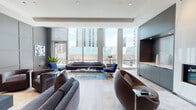 Located on N State St in Chicago's vibrant Gold Coast neighborhood, this elegantly appointed amenity floor delivers a refined blend of modern design and thoughtful functionality. The main lounge area is anchored by floor-to-ceiling windows that flood the space with natural light and frame striking views of the Chicago skyline. Curved brown leather chairs, a sleek gray sofa, and blue accent seating are arranged around glass coffee tables atop light hardwood flooring, creating a welcoming yet polished atmosphere. A dramatic marble-clad fireplace serves as a focal point, complemented by built-in gray cabinetry. The media lounge features a grid-paneled gray accent wall with an integrated flat-screen TV, plush velvet swivel chairs, and curated artwork, offering an intimate setting for relaxed gatherings. The chef-caliber kitchen showcases an expansive marble waterfall island, floor-to-ceiling navy cabinetry, a statement pendant light, and professional-grade appliances. A separate catering pantry is outfitted with warm geometric wallcovering, open wood shelving with decorative accessories, and a long white countertop with under-counter storage. The dining and co-working area provides ample seating at large communal tables alongside flexible smaller groupings, all bathed in natural light from the building's glass curtain wall. Throughout, light hardwood floors, recessed lighting, and a cohesive gray-and-blue palette unify the space.