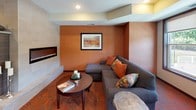 This single-floor communal space is designed with a warm, inviting aesthetic, characterized by rich terracotta and burnt orange accent walls that create a cohesive and energetic atmosphere throughout. The boldly patterned carpet in coordinating orange and gray tones anchors each zone while unifying the open layout.

A central architectural feature — a double-sided tile-clad fireplace column with a mounted flat-screen TV — serves as a natural divider between two distinct lounge areas. One side features a plush gray sectional sofa with colorful accent pillows and an oval coffee table, while the other offers a pair of upholstered plaid armchairs with ottomans and a built-in side table. Both seating areas benefit from recessed lighting overhead.

A separate wing houses a full-size pool table set beneath recessed lights, with a large framed mirror expanding the sense of space. Adjacent to the main lounge, a dining area provides a rectangular table with seating for several guests. An additional TV lounge with a freestanding media console and comfortable seating rounds out the space. Natural light filters in through large windows, enhancing the overall warmth of the environment.