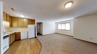 This single-floor space features an open layout that seamlessly connects the kitchen and living area, creating a functional and practical flow. The kitchen is outfitted with warm wood-toned upper and lower cabinetry, a white refrigerator, and hardwood-style flooring that defines the cooking zone. A textured ceiling adds dimension overhead, while flush-mount light fixtures provide consistent illumination throughout.

The living area transitions from the kitchen's hard flooring to neutral wall-to-wall carpeting, offering a comfortable and cohesive feel. White walls keep the interior bright and versatile, while a window along the far wall allows natural light to filter in. Baseboards and clean wall lines contribute to a tidy, straightforward aesthetic.

A doorway visible from the main living space leads to additional areas, suggesting further rooms beyond the open zone. The overall layout is efficient and practical, making good use of the available footprint. The neutral color palette throughout — from the cabinetry to the flooring and walls — provides a clean, move-in-ready atmosphere suited to a variety of furnishing styles.