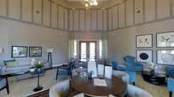 This clubhouse is a well-appointed community gathering space designed to impress and accommodate residents with style and comfort. The interior immediately draws the eye upward with dramatically tall ceilings accentuated by decorative paneling and trim work that lend a refined architectural character to the space. Neutral warm-gray tones on the walls create a sophisticated backdrop that complements the varied furnishings throughout.

The main area is thoughtfully arranged with multiple seating groupings, including a central round dark-wood table surrounded by tufted upholstered chairs, encouraging casual conversation and community interaction. Vibrant teal accent chairs are positioned nearby, adding a pop of color that enlivens the overall palette. Additional seating is arranged closer to the entrance, providing flexible configurations for gatherings of various sizes.

Natural light filters in through large French doors framed by elegant patterned drapery panels, offering a view to the outdoors and connecting the interior to the exterior environment. An overhead chandelier with multiple exposed bulbs provides warm ambient lighting, complementing the floor lamps placed throughout the space. Decorative greenery and area rugs on hardwood-style flooring complete the inviting, community-focused atmosphere of this clubhouse.