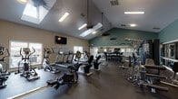 This fitness center is housed within a single-story space distinguished by soaring vaulted ceilings that lend an open, airy quality to the environment. A skylight positioned along the roofline draws in natural daylight, complementing the recessed lighting and suspended ceiling fans that maintain airflow throughout the room. The walls are painted in a neutral beige on the lower half and a deep teal-green on the upper portion, creating a visually defined and energizing atmosphere.

The cardio zone features multiple treadmills, elliptical trainers, upright and recumbent stationary bikes, and a rowing machine, all arranged to allow comfortable spacing between users. Wall-mounted flat-screen televisions are positioned for easy viewing during workouts. A dedicated strength training area includes a multi-station cable machine, a weight bench, and a well-stocked dumbbell rack. Floor-to-ceiling mirrors line one full wall, visually expanding the space and allowing users to monitor their form.

Rubber flooring covers the entire area, providing durability and support underfoot. Large windows on multiple walls offer views to the outdoors, enhancing the sense of openness. Workout reference charts posted on the walls add a practical, informative touch. The overall ambiance is functional, clean, and conducive to focused exercise.