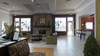 This single-floor common area functions as an inviting lounge or clubhouse gathering space, thoughtfully designed to encourage relaxation and social interaction. The room's most prominent architectural feature is a floor-to-ceiling natural stone fireplace, which serves as a dramatic focal point along the main wall. Decorative accents, including wall-mounted artwork and ceramic vases, are displayed on the fireplace mantel, adding a refined touch to the rustic stonework.

The ceiling is defined by elegant tray detailing with recessed lighting that provides warm, ambient illumination throughout the space. Large windows fitted with horizontal blinds line multiple walls, allowing generous amounts of natural light to filter in while maintaining privacy. A glass-paneled entry door further enhances the brightness of the room and provides a welcoming point of access.

The flooring consists of large-format light-toned ceramic tiles, contributing to the open and airy feel of the space. Comfortable green upholstered sofas and accent seating are arranged around a dark wood coffee table atop a decorative area rug, creating a cozy conversational setting. Tall indoor plants are positioned throughout, adding lively greenery that complements the warm brown accent walls. Overall, the space presents a polished yet comfortable atmosphere suitable for community gatherings.