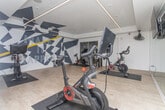 The Spin Room is a purpose-built indoor cycling studio designed to deliver an immersive and high-energy workout environment. The space accommodates multiple Peloton stationary bikes, each positioned on protective floor mats and arranged to provide riders with ample room to train comfortably. Light wood-style flooring runs throughout, offering a clean and modern foundation that contrasts effectively with the room's bold design elements.

The most striking visual feature is the dynamic geometric mural that spans the walls, rendered in dark charcoal, white, and sharp yellow accents. The angular, fragmented pattern creates an energetic atmosphere that reinforces the intensity of a cycling workout. A full-length mirrored wall along one side amplifies the sense of space and allows riders to monitor their form during sessions.

Overhead, recessed linear LED lighting runs along the ceiling perimeter, casting a bright, even illumination across the studio. A ceiling-mounted fan provides additional airflow for comfort during intense sessions. A wall-mounted flat-screen display is also present, likely used for guided workouts or performance tracking. A yellow bench is positioned near the wall, offering a convenient spot for equipment or personal belongings. Glass-panel access connects the spin room to adjacent areas, contributing to a sense of openness within the facility.