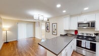 This single-floor residence offers a thoughtfully designed open-concept layout that seamlessly connects the kitchen and living area. The kitchen is a standout feature, showcasing crisp white shaker cabinetry, dark gray quartz countertops, and a central island that provides additional prep and dining space. Stainless steel appliances — including a refrigerator, range, microwave, and dishwasher — complete the modern, functional setup. Recessed lighting and a stylish multi-pendant chandelier illuminate the space effectively.

Warm hardwood flooring flows throughout the main living areas, creating a cohesive and inviting atmosphere. The living area is generously proportioned, with large vertical blinds covering a sliding door that allows natural light to filter in. Neutral, warm-toned walls throughout the residence enhance the bright and airy feel.

The bedroom features plush carpeting and a well-sized window fitted with blinds, offering a quiet and comfortable retreat. The bathroom area includes a vanity with light countertops and gray cabinetry, leading into a full bathroom with a shower/tub combination, toilet, and a window for natural ventilation. Overall, the space presents a clean, modern aesthetic with practical finishes suited for comfortable everyday living.