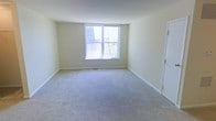 This single-story space presents a clean, minimalist interior with a neutral color palette throughout. The main room is finished with wall-to-wall carpet in a soft gray tone, providing a comfortable and quiet underfoot feel. Pale, light-toned walls contribute to a bright and airy atmosphere, while white baseboards offer a crisp contrast along the perimeter of the room. A large, centrally positioned window serves as a focal point, allowing generous amounts of natural light to fill the space and offering a view of mature trees and surrounding greenery outside. The window is fitted with a simple roller blind, providing privacy and light control as needed. A floor-level heating vent is visible beneath the window, indicating a built-in climate control system. The room's layout is open and versatile, offering ample space for a variety of furniture arrangements. A door visible along the right edge of the room hints at additional areas or storage within the unit. Overall, the space maintains a straightforward, functional design with neutral finishes that provide a blank canvas suitable for personalization.