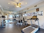 Located at 7000 Cook Rd in Houston, Texas, this fitness center is a well-appointed single-room exercise facility designed to support a well-rounded workout routine. The space features durable dark rubber flooring throughout, crown molding along the ceiling, and a ceiling fan with integrated lighting that promotes air circulation and comfort during workouts. Recessed lighting further brightens the interior, contributing to an energetic and functional atmosphere. A large wall-to-wall mirror runs along one side of the room, paired with a ballet-style barre, visually expanding the space and allowing users to monitor their form. The room is outfitted with a variety of exercise equipment, including a treadmill, an elliptical trainer, a stationary bike, a multi-station cable and weight machine, and an assortment of free weights and dumbbells. Motivational posters adorn the walls, adding an encouraging atmosphere. Natural light enters through windows on multiple walls, including views toward what appears to be an outdoor pool area, accessible via a glass-paneled door. Safety features such as a fire extinguisher and sanitation dispensers are also present. The overall design is clean, practical, and purpose-built for fitness.