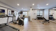 Located at 115 Hutchinson Ave in Columbus, Ohio, this well-appointed fitness center offers a practical and comfortable environment for exercise. The single-floor layout makes efficient use of its space, providing a dedicated area for both cardio and strength training activities.

The main workout area features light wood-style flooring that lends a clean, modern aesthetic throughout. Recessed ceiling lighting ensures the room is well-lit, while multiple windows with horizontal blinds allow natural light to filter in, creating a bright and inviting atmosphere. Structural columns are integrated into the layout, subtly dividing the space without disrupting flow.

Cardio equipment includes multiple treadmills, an elliptical, a recumbent bike, and a stationary cycle — each equipped with individual monitors for entertainment or workout tracking. A wall-mounted flat-screen TV provides an additional viewing option for users. A rack of dumbbells and adjustable weight benches support strength training needs, while a body weight scale is available near the perimeter. A water dispenser station, storage cabinet, and open shelving unit provide convenient amenities within the space.

A utility room is also present, likely serving maintenance or storage purposes. Framed artwork decorates the walls, contributing to a polished, welcoming environment. Overall, this fitness center is thoughtfully equipped to support a well-rounded workout routine.